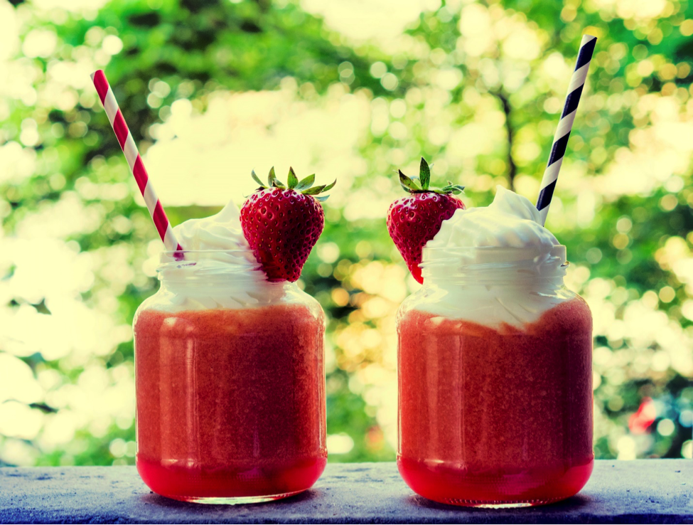 Two containers of a strawberry drink topped with a strawberry, whipped cream and straws