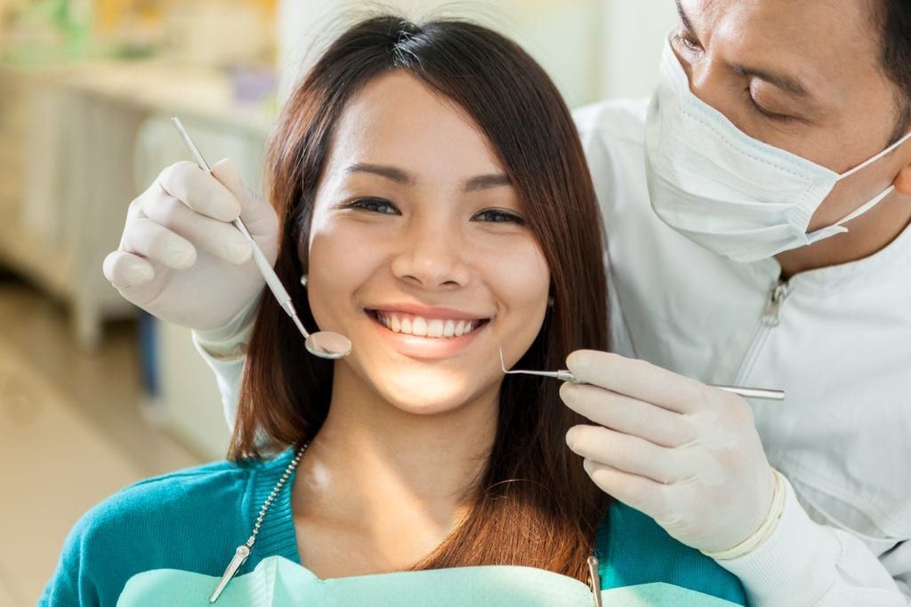 male dentist holding a mirror mouth and curette while female patient smiles