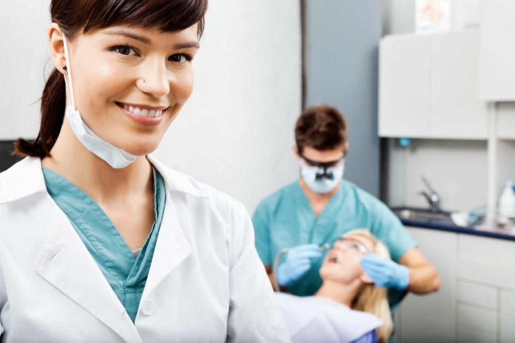 female dentist smiling with a male dentist cleaning a female patient teeth in the backfground