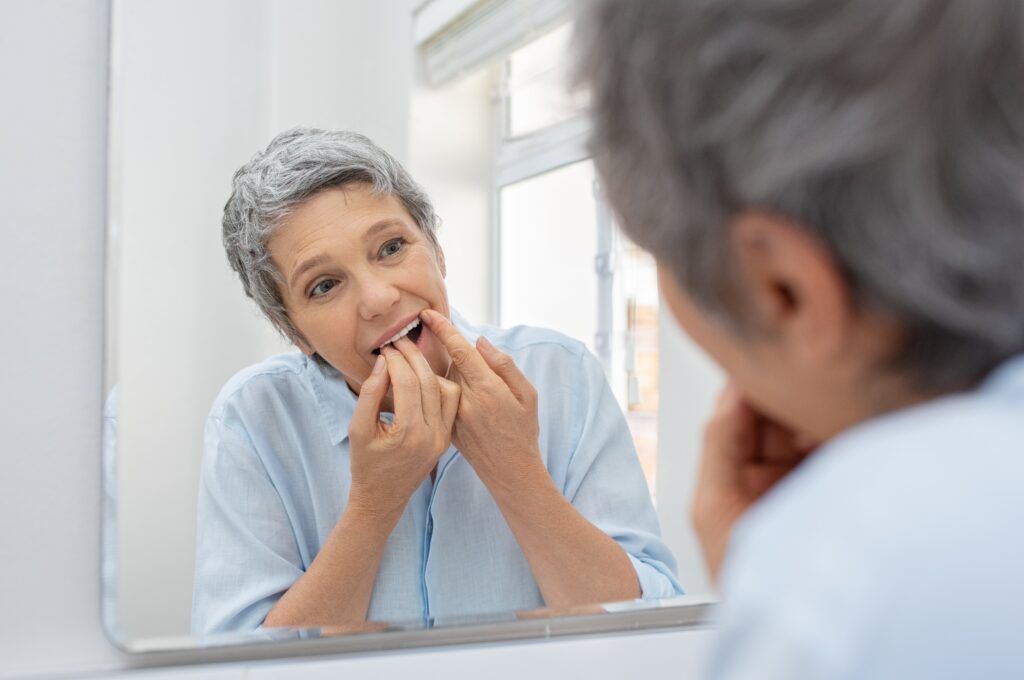older woman flossing her teeth in mirror