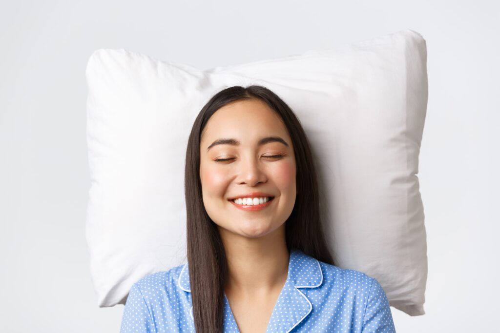 Smiling dreamy and beautiful asian female lying in bed on pillow, wearing blue pajamas, close eyes and grinning, daydreaming or sleeping at night, imaging something cute, white background